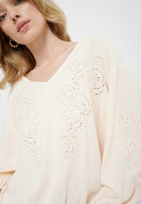 Lichtbeige blouse met een V-hals, voorzien van delicate borduursels en uitsnijdingen op de schouders en zijkanten. Gemaakt van zachte stof.