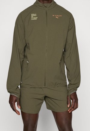 Mann trägt olivgrüne Sportjacke und Shorts mit den Logos „World Series of Fitness Racing“ und „HYROX“, steht vor einfarbigem Hintergrund.
