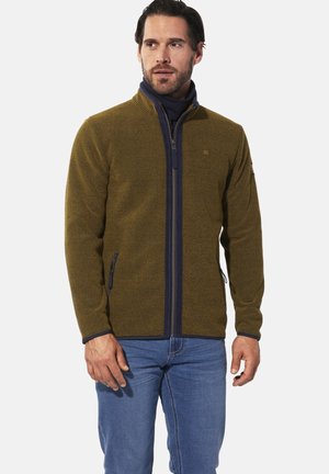 Olivgrüne Fleecejacke mit Reißverschluss und strukturiertem Muster, navyfarbene Akzente und Seitentaschen, kombiniert mit blauen Jeans.
