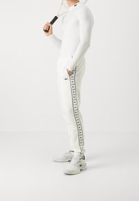 Bílá atletická košile s dlouhým rukávem v kombinaci s bílými tepláky se černými bočními pruhy a logem Lacoste. Šedé tenisky dotvářejí vzhled.