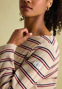 Beige gestreifter Pullover mit kastanienbraunen und marineblauen Linien, gerippten Bündchen und einem Logo-Patch auf der linken Schulter. Glatte Baumwolltextur.