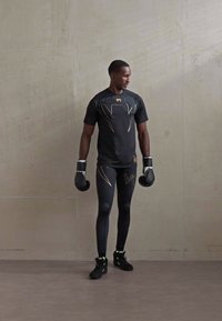 Hombre con guantes de boxeo negros, una camiseta deportiva negra con un diseño de serpiente dorada, mallas negras y zapatos negros, de pie contra una pared lisa.