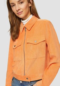 Oranje corduroy jas met een rits, twee borstzakken en knoopaccenten; heeft een kraag en ribbeltextuur.