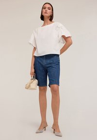 Top bianco a maniche corte con dettagli arricciati, abbinato a pantaloni bermuda in denim blu. Il modello tiene in mano una piccola borsa color crema e indossa mule grigie con tacco.
