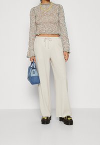 Osoba v béžovém krátkém svetru, krémových širokých kalhotách, černých platformových botách, s žlutým náhrdelníkem a modrou patchwork kabelkou.