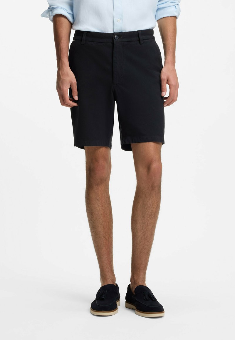 Homme portant un short noir sur mesure, une chemise bleu clair à manches longues avec les manches retroussées, et des mocassins noirs à pampilles, debout sur fond blanc.