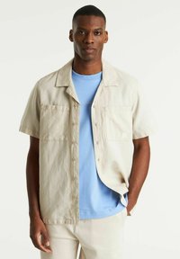 Beige korte mouwen overhemd met twee zakken aan de voorkant, gedragen over een lichtblauwe ronde hals t-shirt. Eenvoudig ontwerp, lichte stof, casual pasvorm.