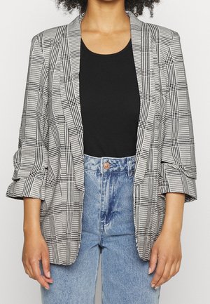 Kobieta ubrana w czarny top z dekoltem w łódkę, kraciastą marynarkę z marszczonymi rękawami i jasnoniebieskie, wysokowalne dżinsy, stojąca na białym tle.