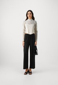 Balta nėriniuota ilgos rankovės crop top palaidinė su sudėtingais raštais, derinama su juodomis klostuotomis plataus kirpimo kelnėmis. Modelis laiko juodą rankinę.