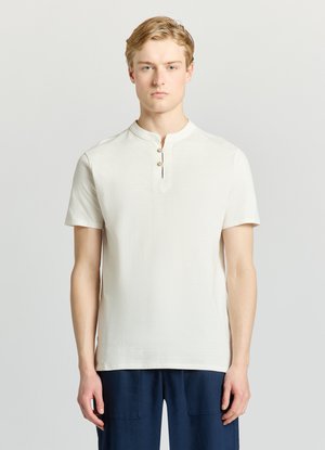 Giovane uomo che indossa una maglia henley bianca a maniche corte con tre bottoni e pantaloni blu navy, in piedi contro uno sfondo grigio chiaro.