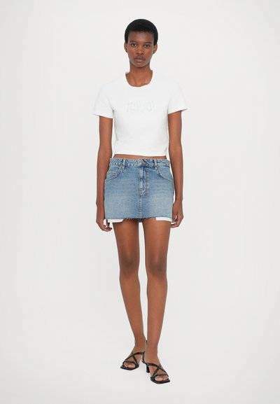 Tricou alb scurt, cu logo texturat, asociat cu o fustă mini din denim albastru, cu tiv desfăcut și buzunare. Sandale negre cu barete.