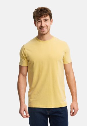 Uomo sorridente con capelli ricci che indossa una maglietta gialla a maniche corte semplice e jeans blu scuro, in piedi contro uno sfondo bianco.