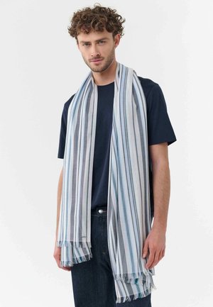 Man wearing navy t-shirt and dark jeans with light blue, white, and gray striped scarf draped over shoulders.