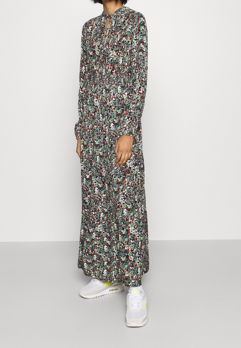 Personne portant une robe fleurie à manches longues, longueur cheville, avec une taille smockée et des baskets blanches avec des accents jaune fluo.