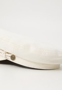 Beret en coton ivoire avec une texture lisse, présentant un accent tressé et un bouton décoratif sur le côté.