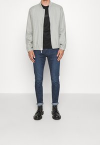 Ljusgrå bomberjacka med ståkrage, svart t-shirt, mörkblå slim jeans och svarta snörstövlar, visad framifrån mot en enfärgad bakgrund.