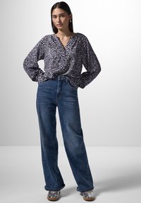 Langärmlige Bluse im schwarz-weißen abstrakten Muster, kombiniert mit hochtaillierten, weit geschnittenen blauen Jeans und silbernen metallic Schuhen.