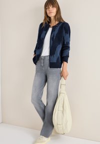 Marineblauer, flauschiger Pullover mit strukturiertem Design, graue Jeans und eine cremefarbene, lässige Tasche. Das Modell trägt ein weißes Oberteil und neutrale Sneakers.