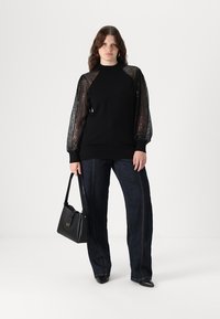 Μαύρη μπλούζα με διάφανα, παγιέτα μανίκια; συνδυασμένη με σκούρα τζιν παντελόνια; μοντέλο κρατώντας μια μαύρη τσάντα, όρθιο σε λευκό φόντο.