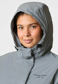 Grijze waterdichte jas met een capuchon, voorzien van een hoge kraag, zwarte hardware en een logo op de borst. Gladde textuur, lichtgewicht ontwerp.