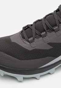 Zapatilla deportiva negra con parte superior de malla texturizada, diseño aerodinámico, cordones y suela de goma gris claro con picos de tracción robustos.