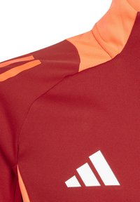 Rotes Sporttrikot mit orangefarbenen Akzenten und Mesh-Struktur. Es hat drei weiße Streifen und ein auffälliges weißes Logo auf der Vorderseite.