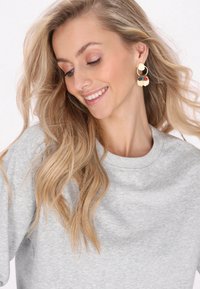 Orecchini circolari dorati con accenti neri, rossi e verdi, caratterizzati da elementi a petalo sovrapposti; modella che indossa un felpa grigia chiara.