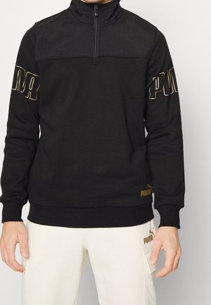 Mann trägt schwarzen Viertel-Zip-Sweatshirt mit goldenem „PUMA“-Schriftzug auf den Ärmeln und weiße Hose mit goldenem Puma-Logo auf der Tasche.