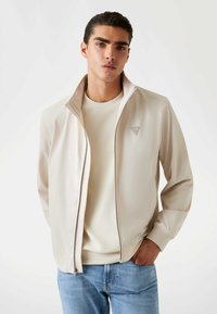 Jonge man met donker haar, gekleed in een beige zip-jack over een crème trui en lichtblauwe jeans, staand tegen een witte achtergrond.