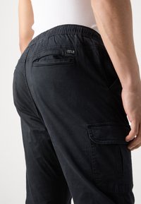 Homme portant un pantalon cargo noir avec une ceinture élastique et plusieurs poches, main reposant près de la poche cargo latérale.