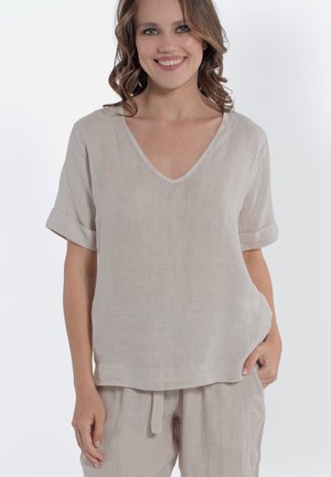 Donna sorridente con capelli castani mossi che indossa una maglietta beige in lino a maniche corte con scollo a V e pantaloni abbinati con coulisse, posando con la mano in tasca.