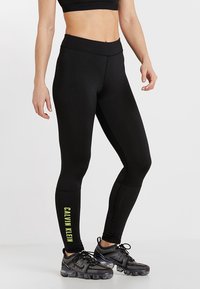 Kobieta w czarnych legginsach Calvin Klein z żółtym logo na lewej łydce oraz czarnych butach sportowych z przezroczystymi podeszwami.