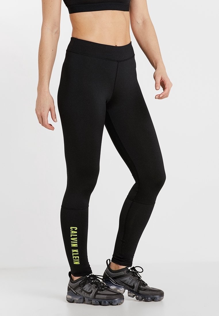 Kobieta w czarnych legginsach Calvin Klein z żółtym logo na lewej łydce oraz czarnych butach sportowych z przezroczystymi podeszwami.