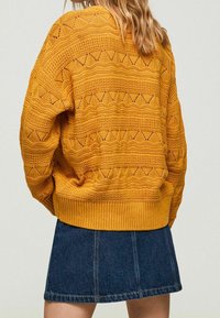 Gestrickter Pullover in Senfgelb mit einem strukturierten Muster aus Zickzack- und Lochstrickdesigns, kombiniert mit einem Jeans-Minirock.