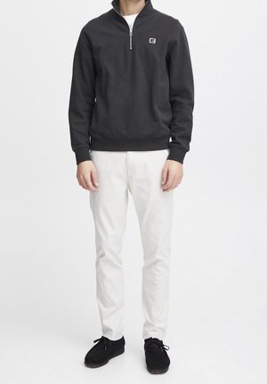 Mężczyzna ubrany w czarną bluzę zapinaną na zamek do połowy długości z logo na piersi, białe spodnie i czarne sznurowane buty, stojący na białym tle.