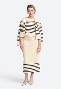 Ensemble tricot crème avec des rayures marines, comprenant un haut court, une longue jupe et une veste assortie. Des chaussures à bout pointu complètent le look.