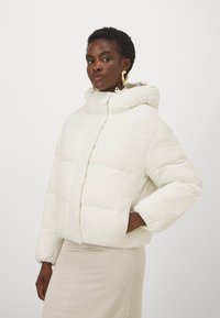 Giacca a vento bianca con cappuccio, design trapuntato, polsini elasticizzati e zip frontale. Texture liscia e taglio corto e ampio.