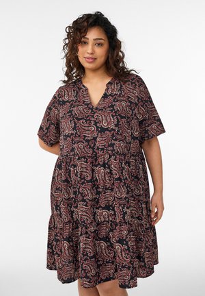 Krulligharige vrouw die een losse zwarte jurk met rood paisley patroon draagt, staand tegen een eenvoudige witte achtergrond.