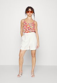 Camisola de alças finas com estampa floral, combinada com calções beije de cintura alta. O modelo usa óculos de sol laranja e sandálias com tiras.