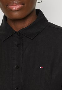 Černá košile na knoflíky s klasickým límečkem, s malým vyšítým logem v červené, bílé a modré na levé hrudi. Texturovaná látka.