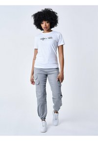 Witte kortekous T-shirt met zwarte tekst, gecombineerd met grijze cargobroek met zijzakken en elastische boorden, aangevuld met witte sneakers.