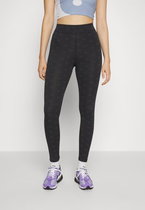 Femme portant des leggings noirs à motifs, des chaussettes blanches et des chaussures de sport violettes et noires, debout devant un fond clair et uni.