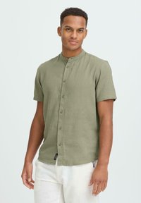 Chemise en lin vert olive à manches courtes avec fermeture à boutons sur le devant et col mandarin. Dotée d'une coupe droite et d'une étiquette à l'ourlet.
