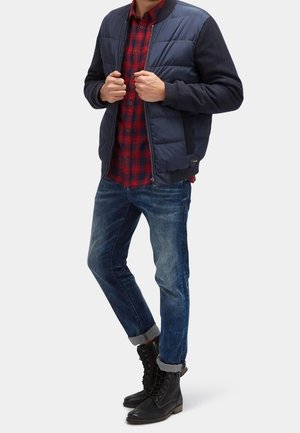 Mężczyzna w granatowej bomberce na czerwonej koszuli w kratę, z podwiniętymi niebieskimi dżinsami i czarnymi sznurowanymi butami, stojący, poprawiający kurtkę.