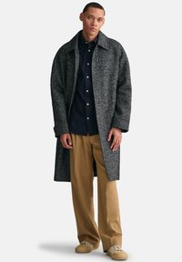 Grijze textuurjas met een brede kraag, lange mouwen en zijsplitten, gelaagd over een donker shirt en tanbroeken, gecombineerd met beige sneakers.