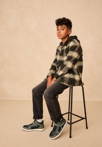 Chemise en flanelle à carreaux vert et noir, portée sur un sweat à capuche sombre. Jean foncé et baskets montantes vert/noir complètent le look.