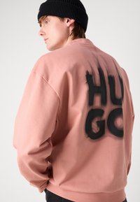 Rózsaszín, laza fazonú pulóver, melynek hátán egy nagy fekete, spray-vel festett "HUGO" felirat található. Fekete kötött sapkával viselve.
