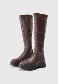 Bottes en cuir marron, arrivant au genou, avec une fermeture éclair sur le côté, un bout rond et une épaisse semelle en caoutchouc noir avec un motif texturé.