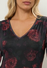 Top negro con escote en V, que presenta grandes patrones florales en burdeos y hojas verdes. Tela texturizada con detalles fruncidos en las costuras de los hombros.