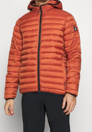 Homme portant une veste à capuche matelassée orange avec fermeture éclair et un pantalon noir, debout devant un fond clair uni.